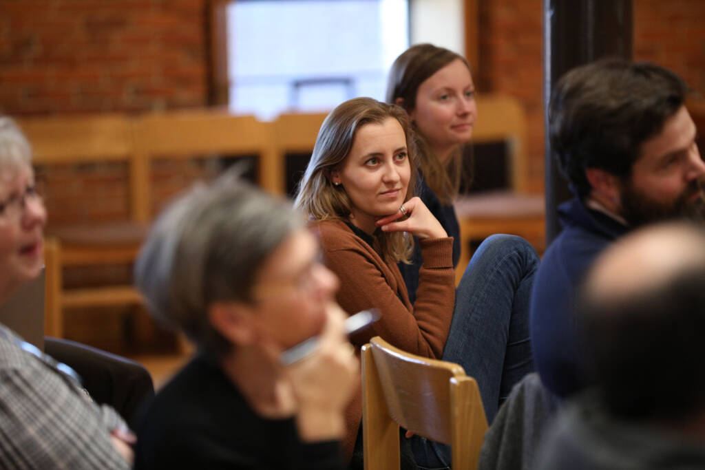 A woman in a brown sweater sits among a group of people in a room, resting her chin on her hand and listening attentively. Other attendees are slightly out of focus around her.