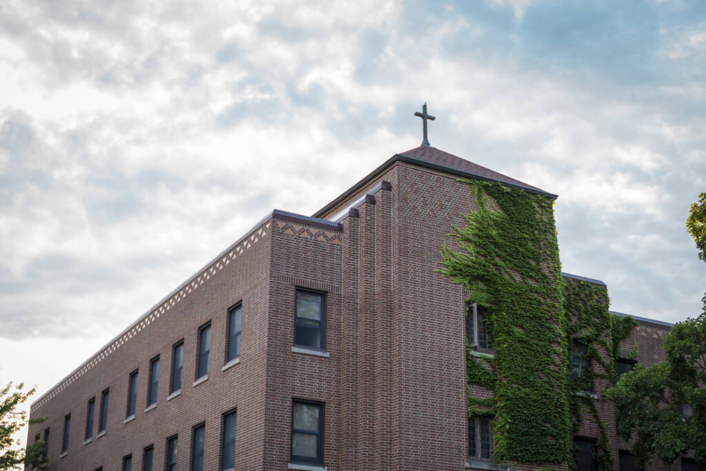 A brick building with a cross on the roof and green ivy climbing one side, set against a partly cloudy sky.