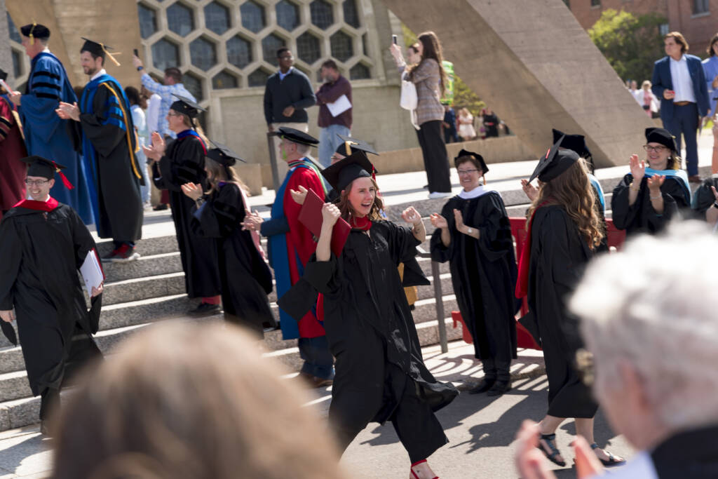 A jubilant graduate in a black cap and gown walks outside, celebrating with fists raised as others clap and cheer around her during a graduation ceremony.