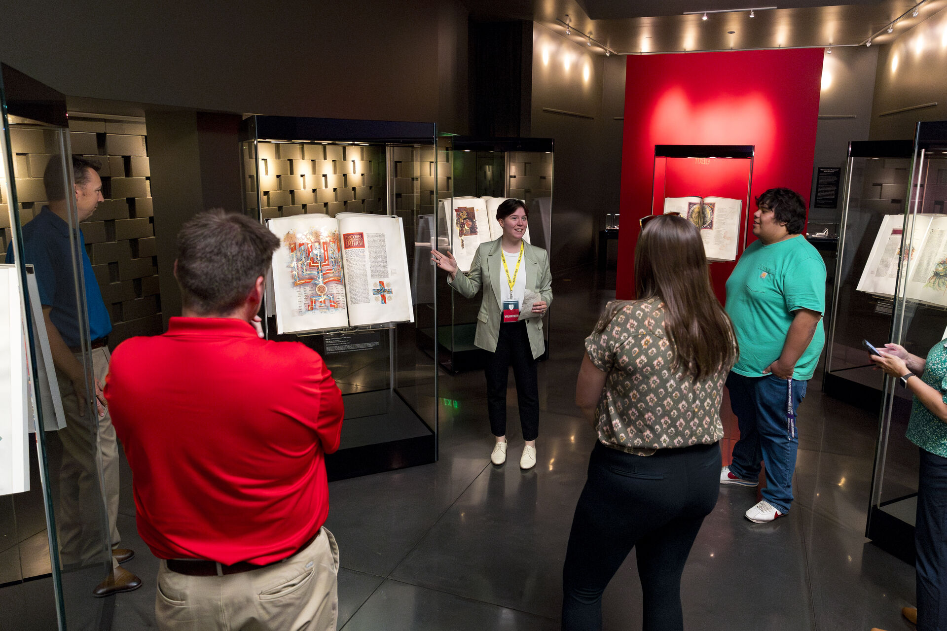 A woman gives a tour to four people in a dimly lit museum exhibit featuring illuminated manuscripts in glass cases, with a large red heart design on the back wall.