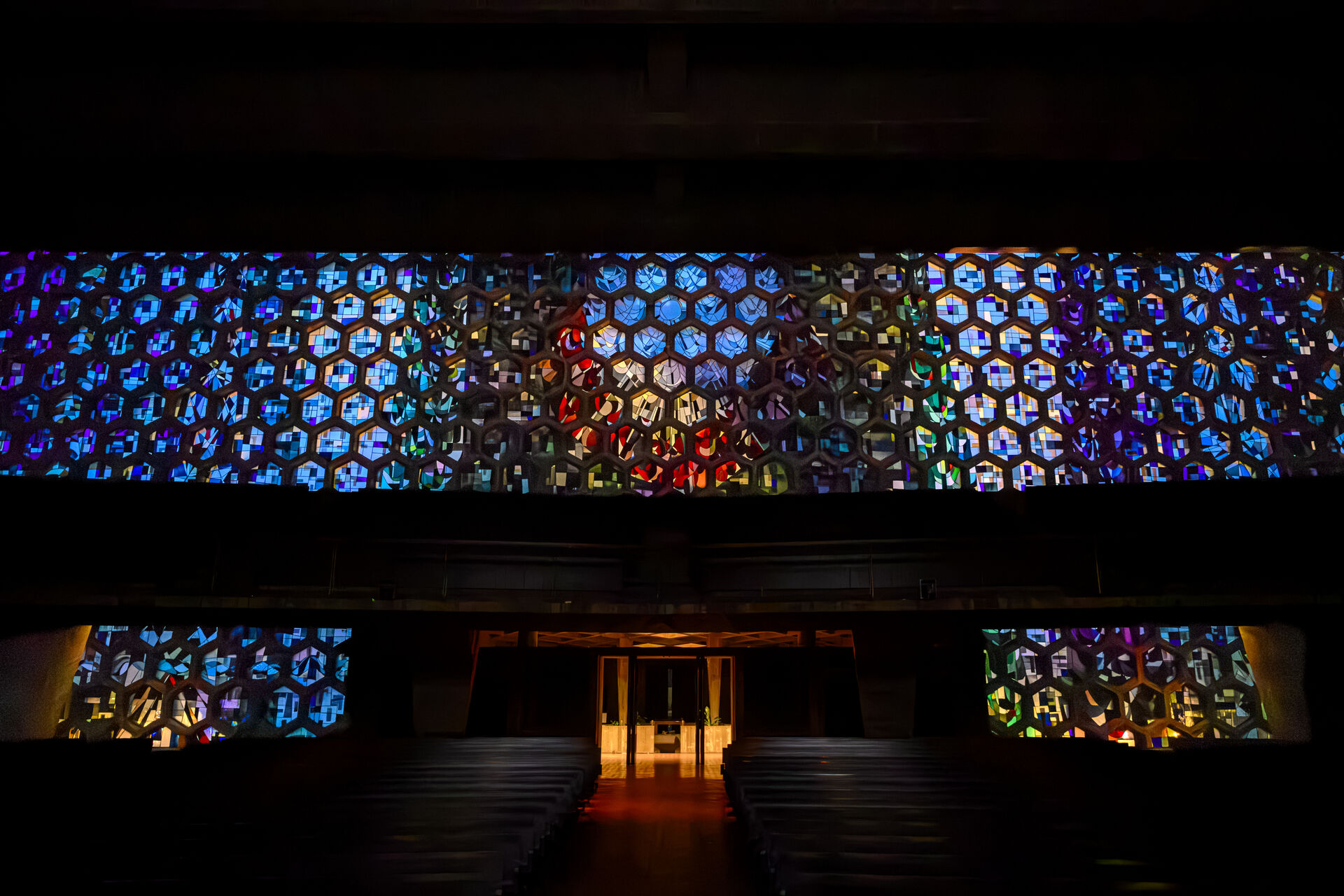 A dimly lit church interior with rows of pews, illuminated by colorful geometric stained glass windows covering the upper walls, and a softly glowing altar at the center in the background.