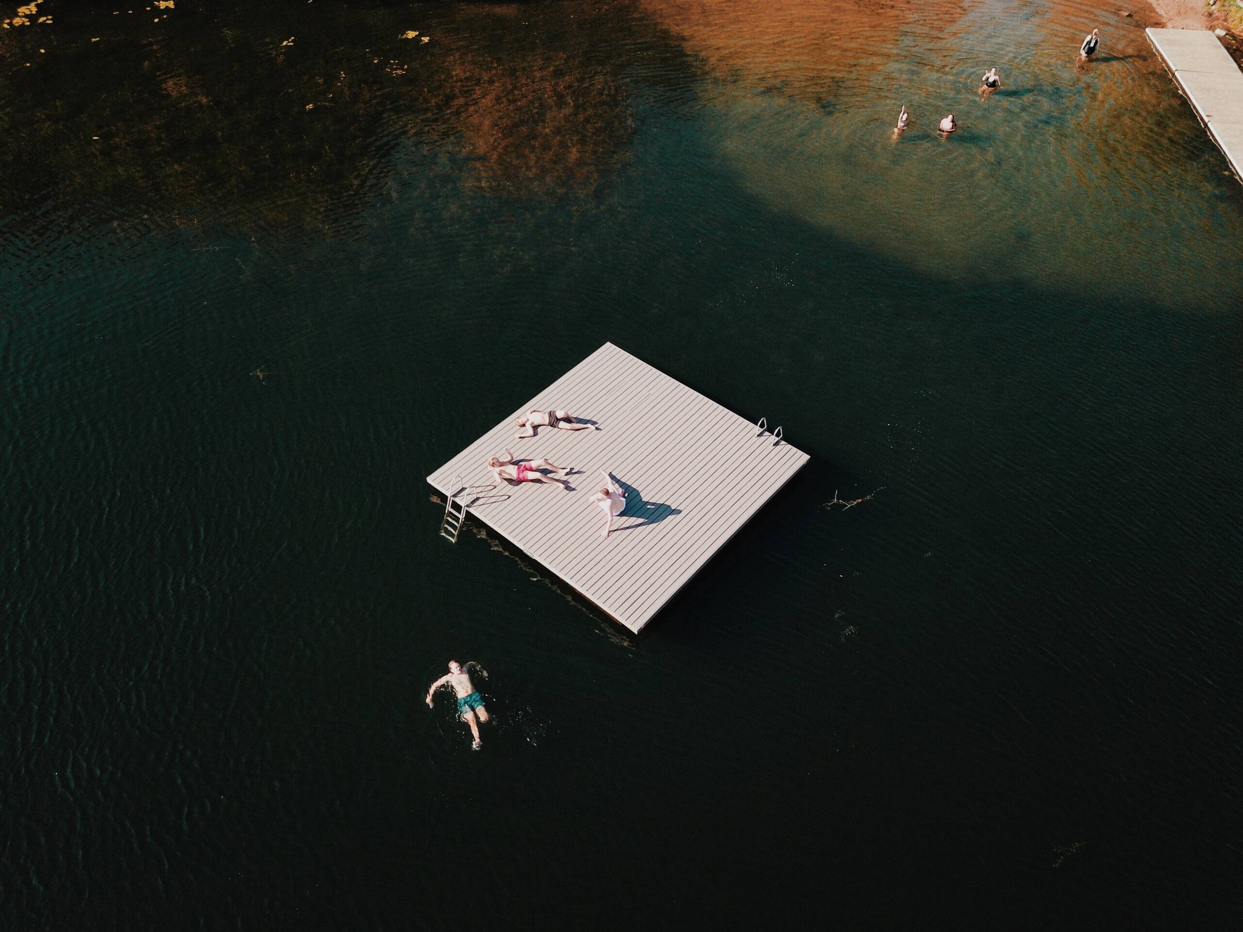 Aerial view of people sunbathing on a floating dock in a lake, with others swimming nearby and a few by a distant pier. The water is dark, reflecting the surroundings and the dock casts a shadow.