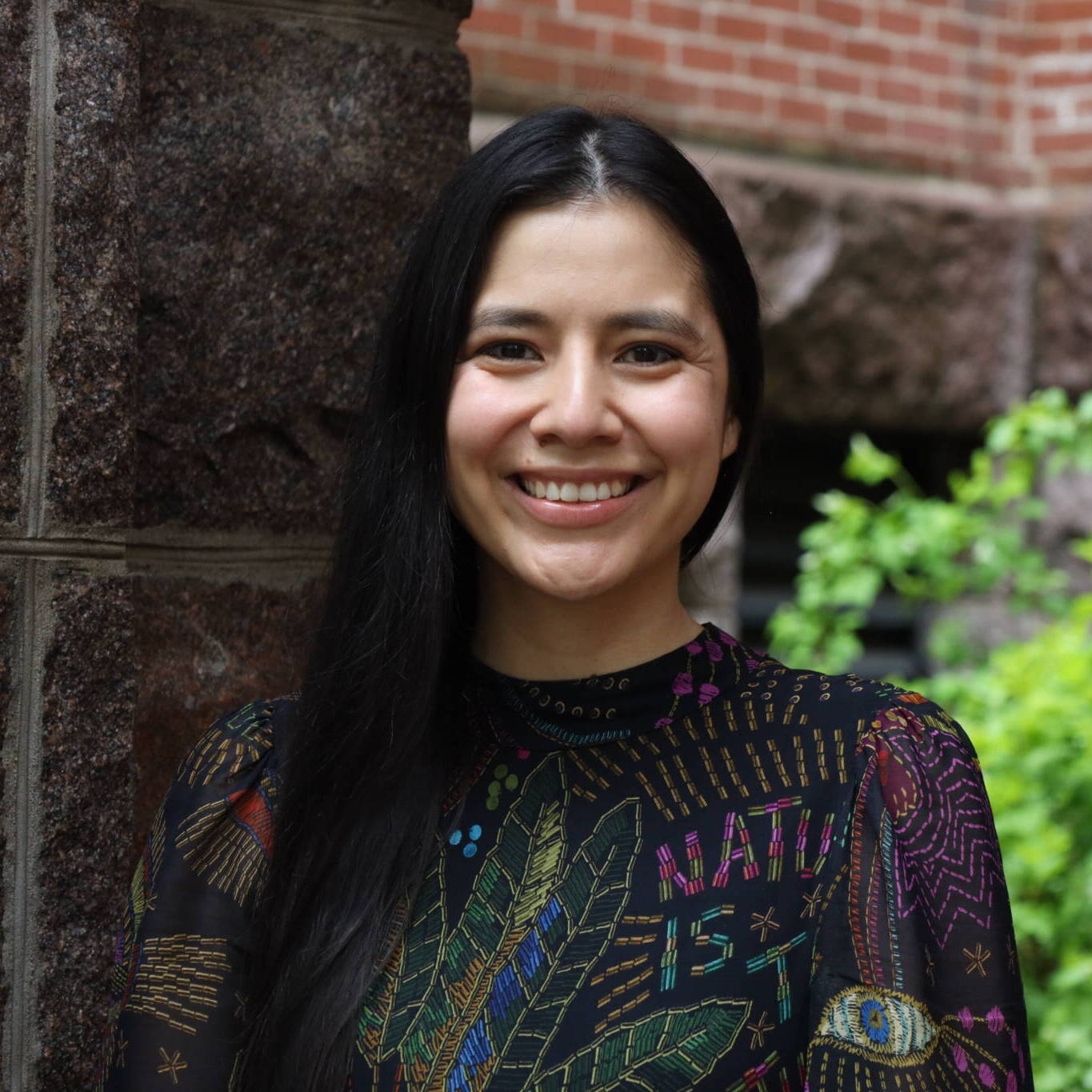 A woman with long dark hair smiles while standing outside near a stone wall and green bushes, with a brick building in the background. She is wearing a colorful patterned top.