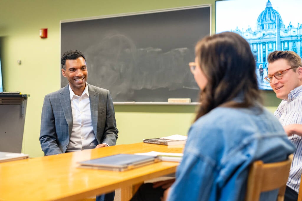 A man in a suit sits at a table, smiling and talking with two people who are seated with notebooks, in a classroom with a chalkboard and a screen displaying a historic building.