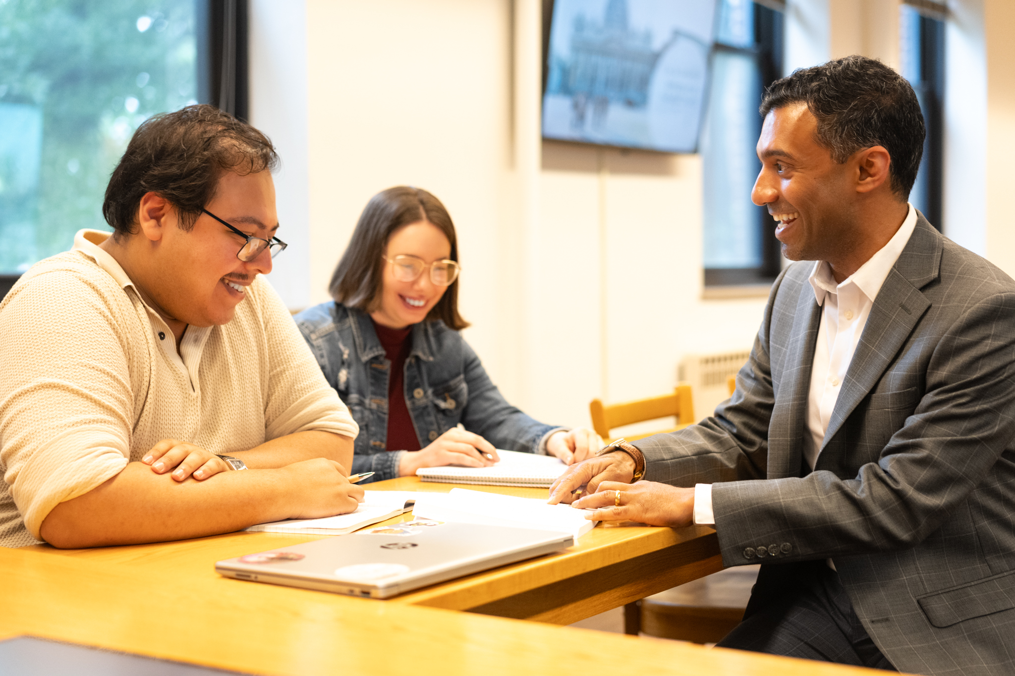 Three people sit at a table in a bright room, smiling and talking while working on papers and a laptop. One person wears a suit, while the other two are dressed casually.