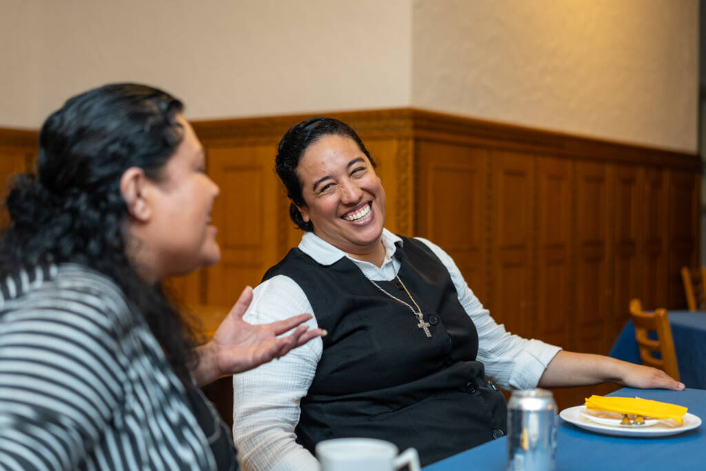 Two women sit at a table, smiling and conversing. One wears a striped jacket, the other a vest with a cross necklace, smiling brightly. Plates, cups, and a soda can are on the table in a warmly lit room.