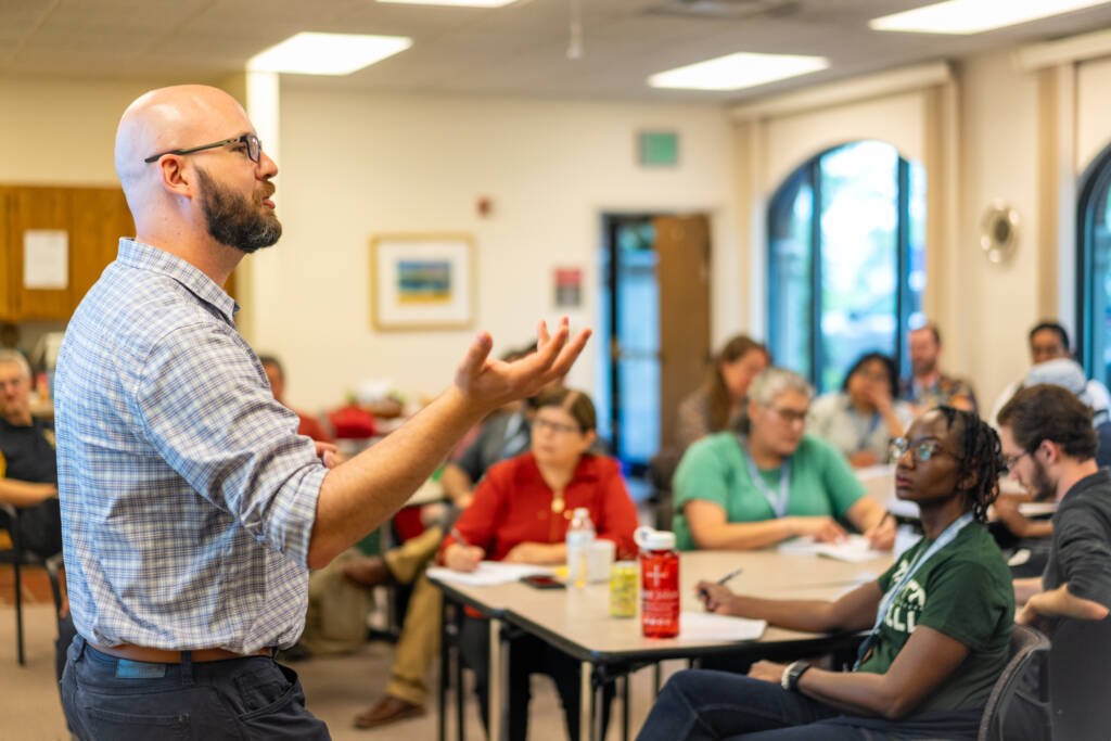 A man with a beard and glasses speaks to a group of people seated at tables, some taking notes and listening attentively, in a bright, classroom-like setting.