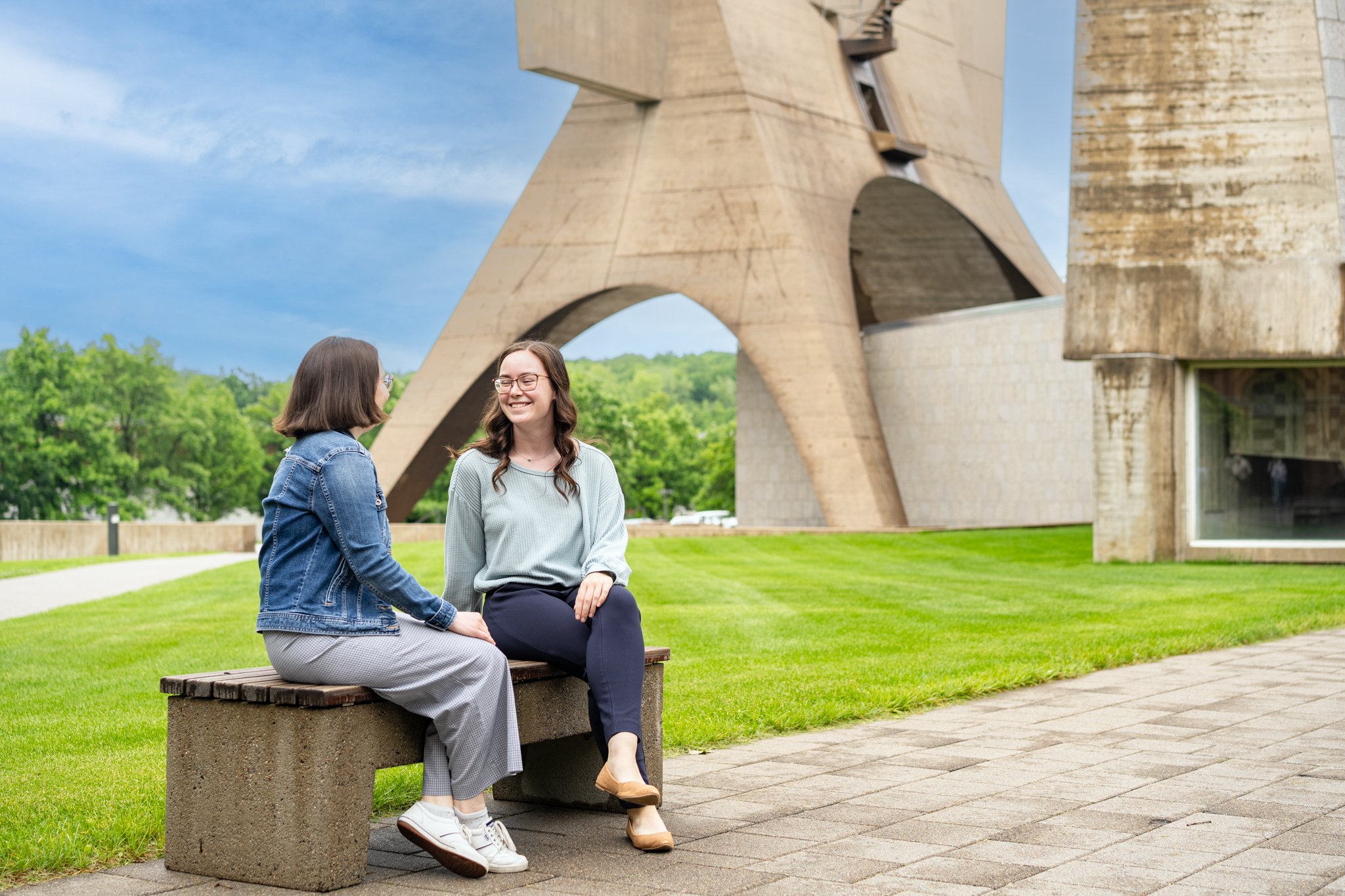 Two women sit on a bench, smiling and talking, in front of a large modern concrete structure with arches. The scene is outdoors, with green grass and trees under a blue sky.