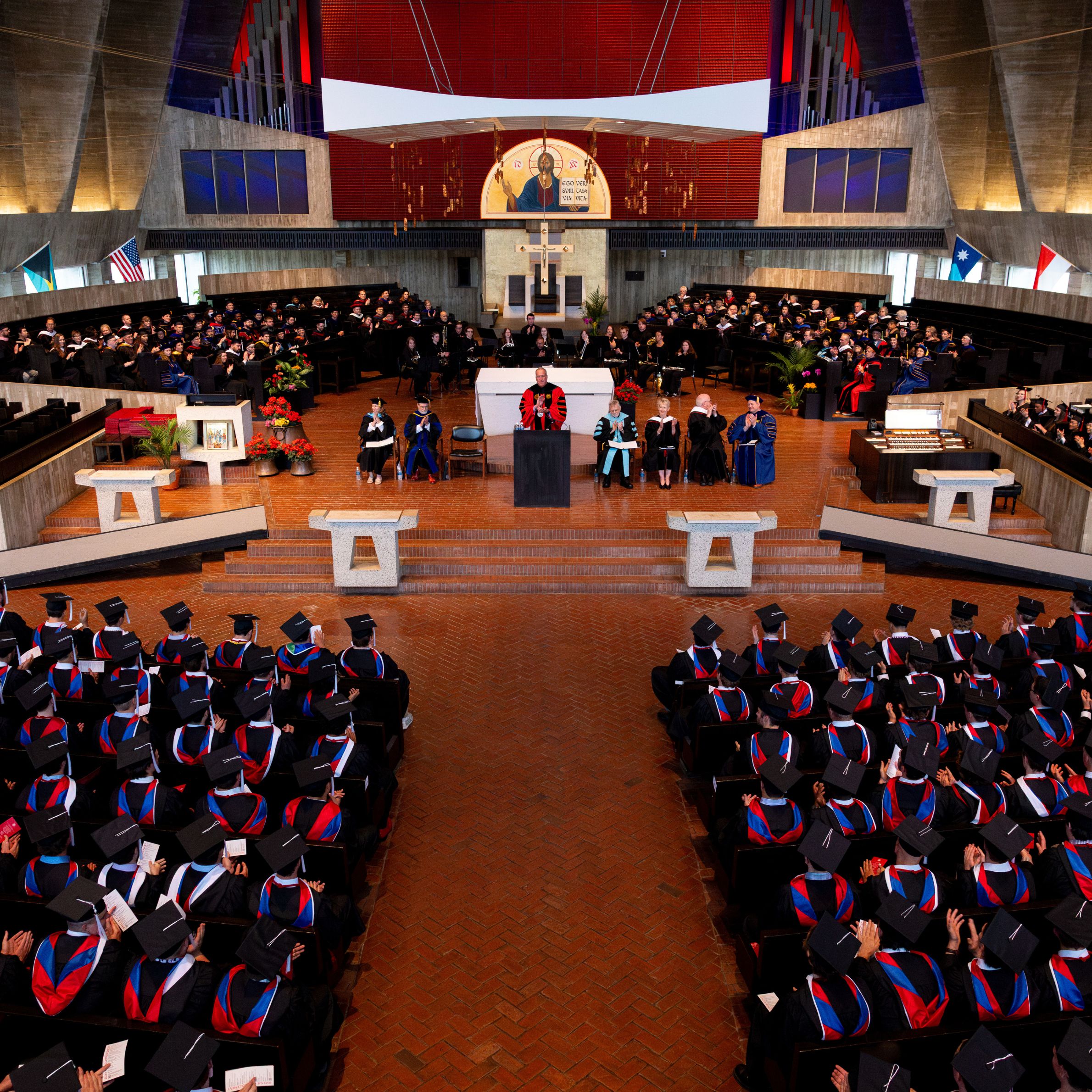 A graduation ceremony inside a large church, showing graduates in caps and gowns seated, faculty on stage, and a speaker addressing the audience from a podium. Flags and religious symbols are visible in the background.