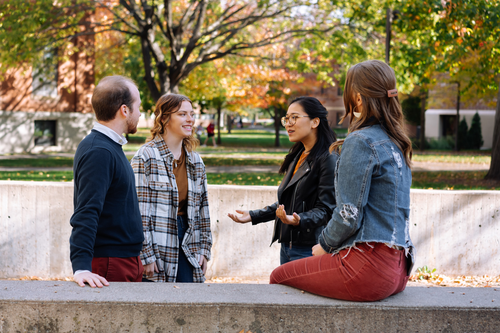 Four young adults, three women and one man, are talking and smiling together outdoors on a sunny autumn day, with colorful trees and a building in the background. One woman sits on a low wall while the others stand.