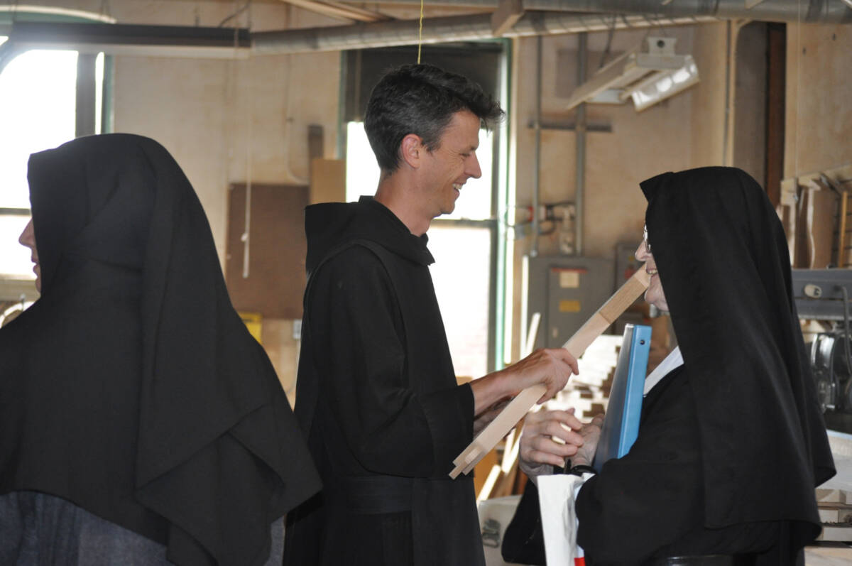 Three people wearing black religious habits stand indoors, two facing each other holding a piece of wood and smiling, while the third stands to the left, mostly turned away from the camera.