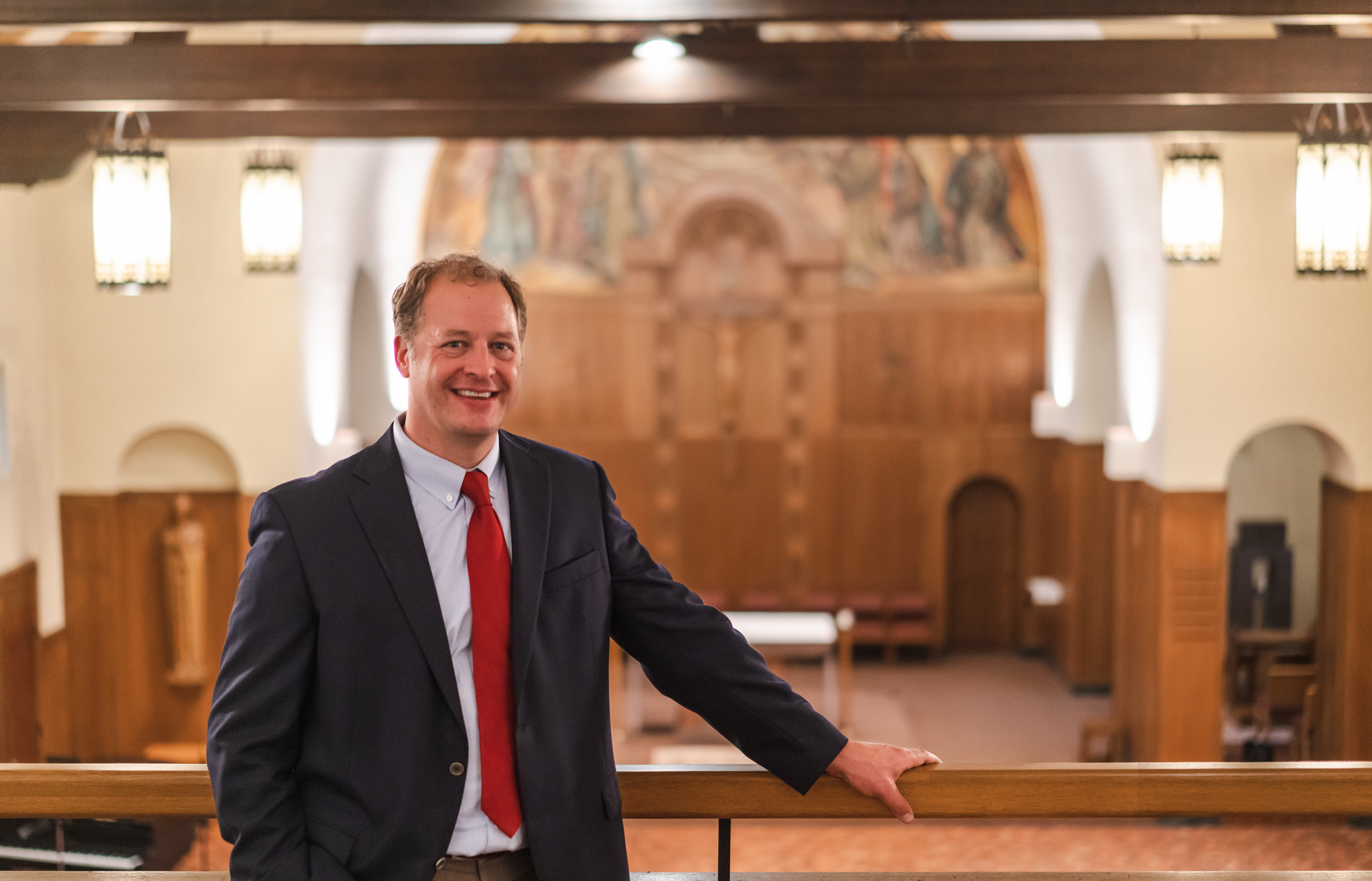 A man in a suit and red tie smiles while leaning on a railing inside a church with wooden interiors and religious artwork on the wall in the background.