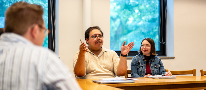 Three people sit at a wooden table in a classroom. One person in the center gestures while speaking, while another listens and smiles. A third person is slightly out of focus in the foreground. Windows show trees outside.