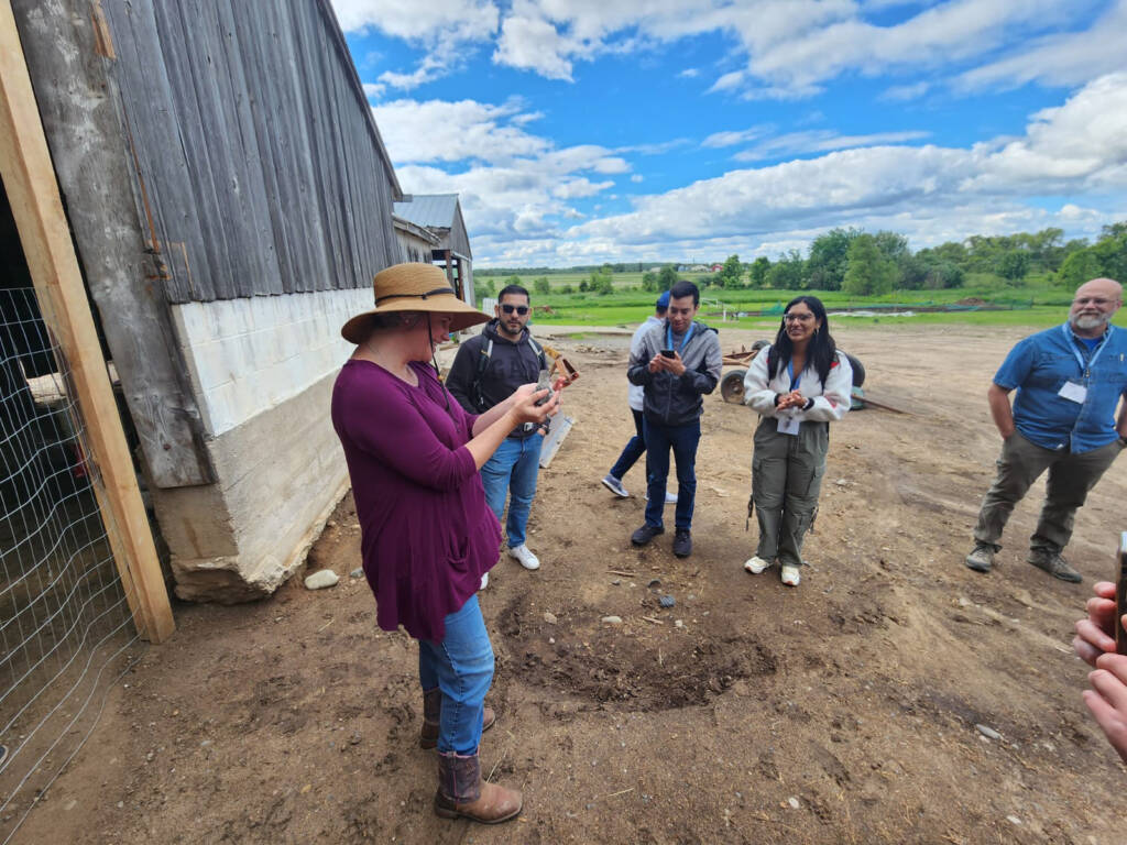 A woman in a sunhat stands on a farm, holding up an object and speaking to a group of people who are attentively listening. The group stands on dirt near a wooden building, with green fields and a cloudy blue sky in the background.
