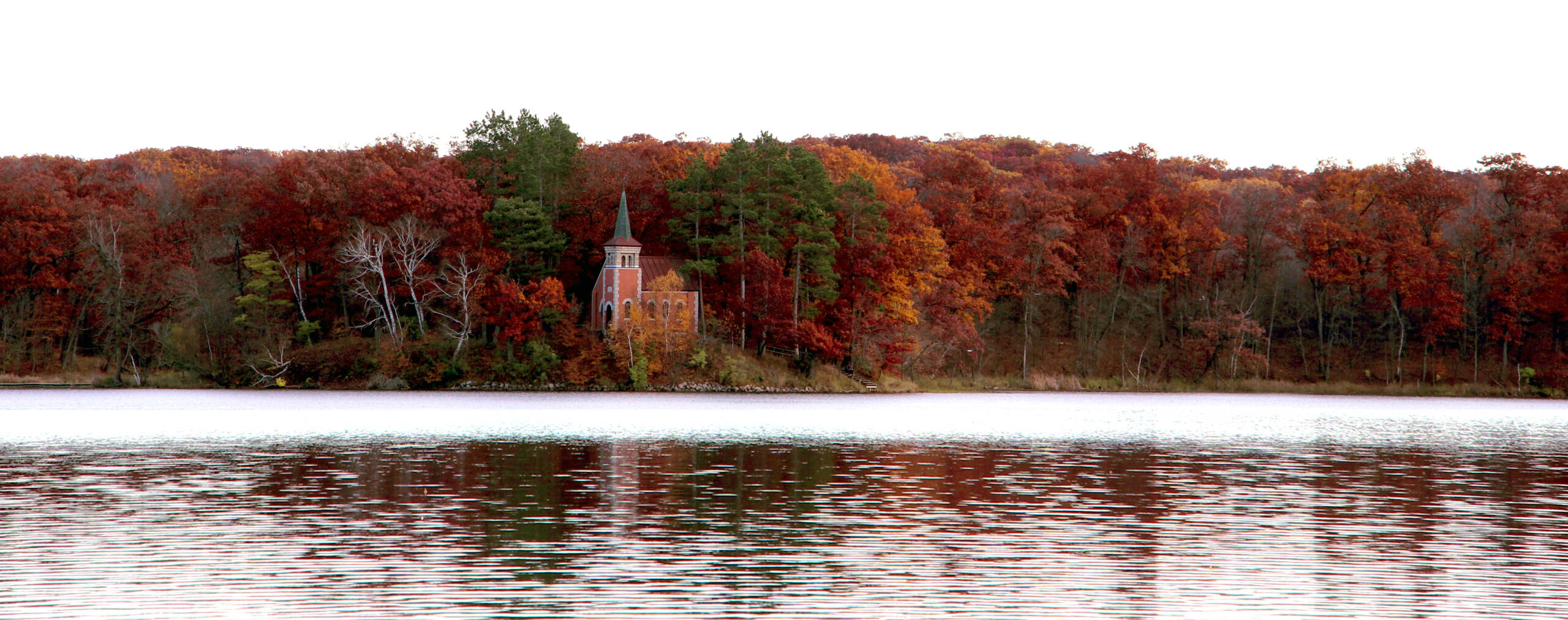 A small brick church with a steeple stands surrounded by colorful autumn trees on the shore of a calm lake, with the foliage reflecting in the water under an overcast sky.