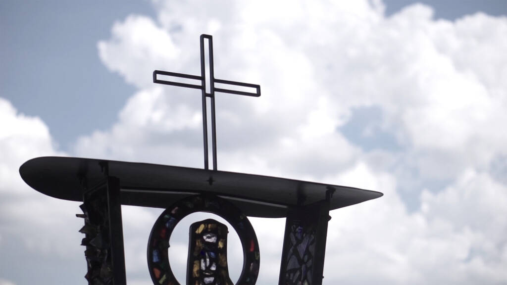 A metal cross stands atop a modern sculpture with stained glass elements, set against a cloudy sky.