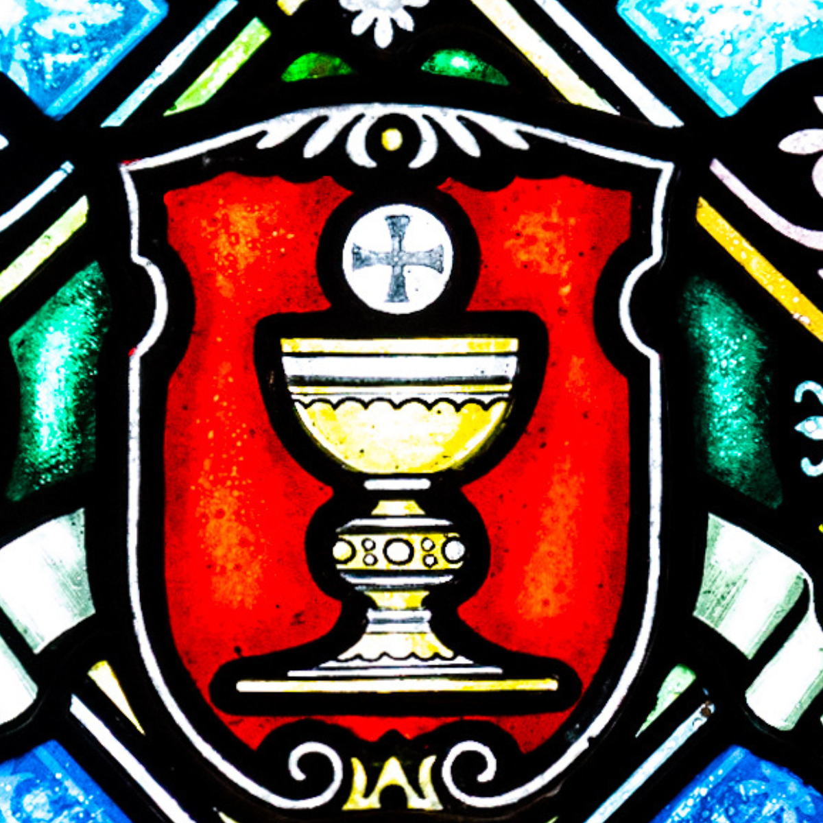 A stained glass window depicts a gold chalice holding a white host with a cross, set against a red shield background with decorative blue, green, and white details.