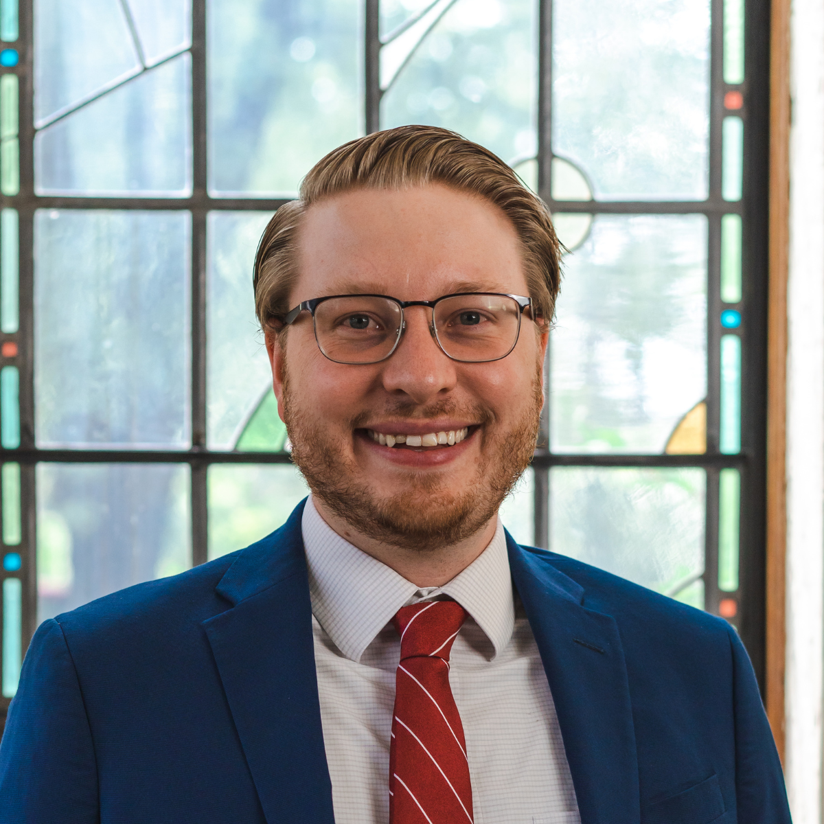 A man with glasses and blond hair, wearing a blue suit, white shirt, and red striped tie, smiles while standing in front of a stained glass window.