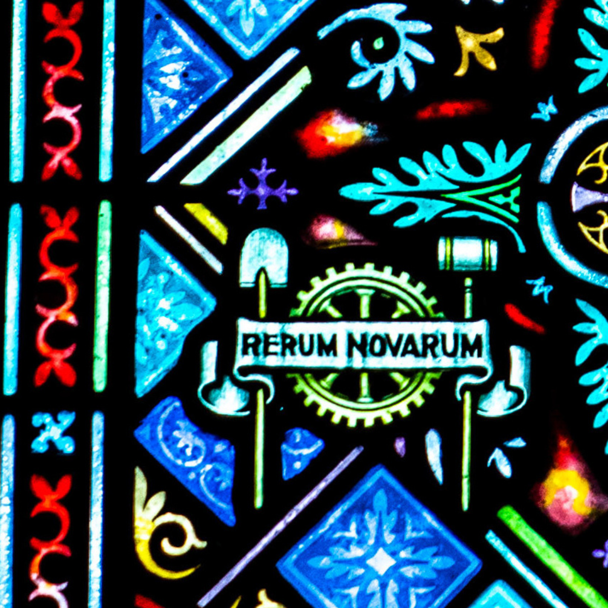Colorful stained glass window with decorative patterns, featuring a banner that reads "Rerum Novarum" held by two hands in front of a gear, with green leaves and various geometric designs in the background.