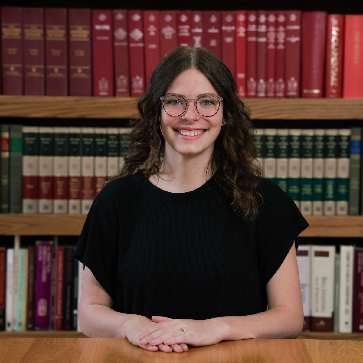 A woman with wavy brown hair and glasses, wearing a black shirt, sits at a wooden table with her hands folded. She is smiling and there are shelves of colorful books behind her.