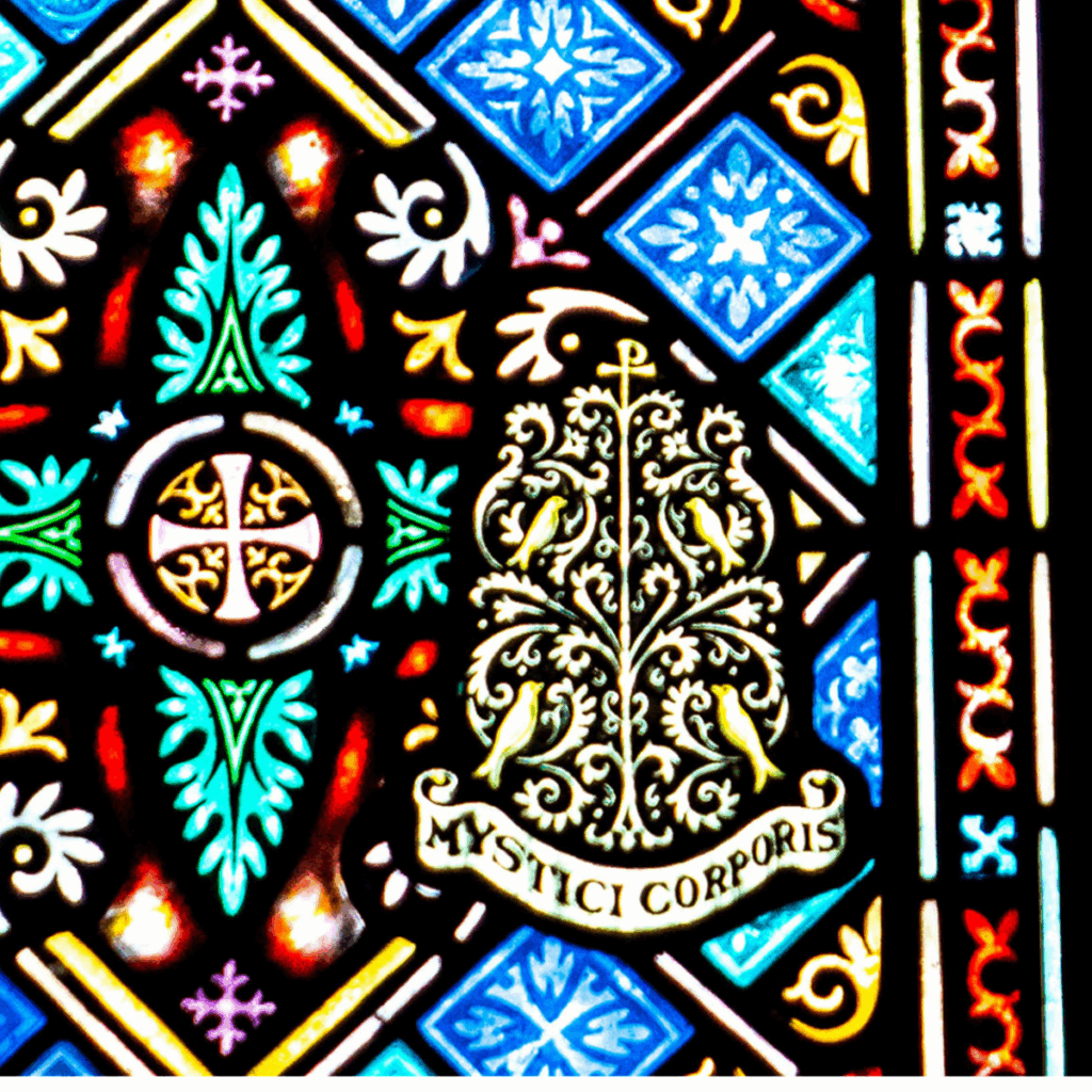 Colorful stained glass window featuring ornate geometric and floral patterns, with a leafy design and a banner below reading "MYSTICI CORPORIS" in gold letters.