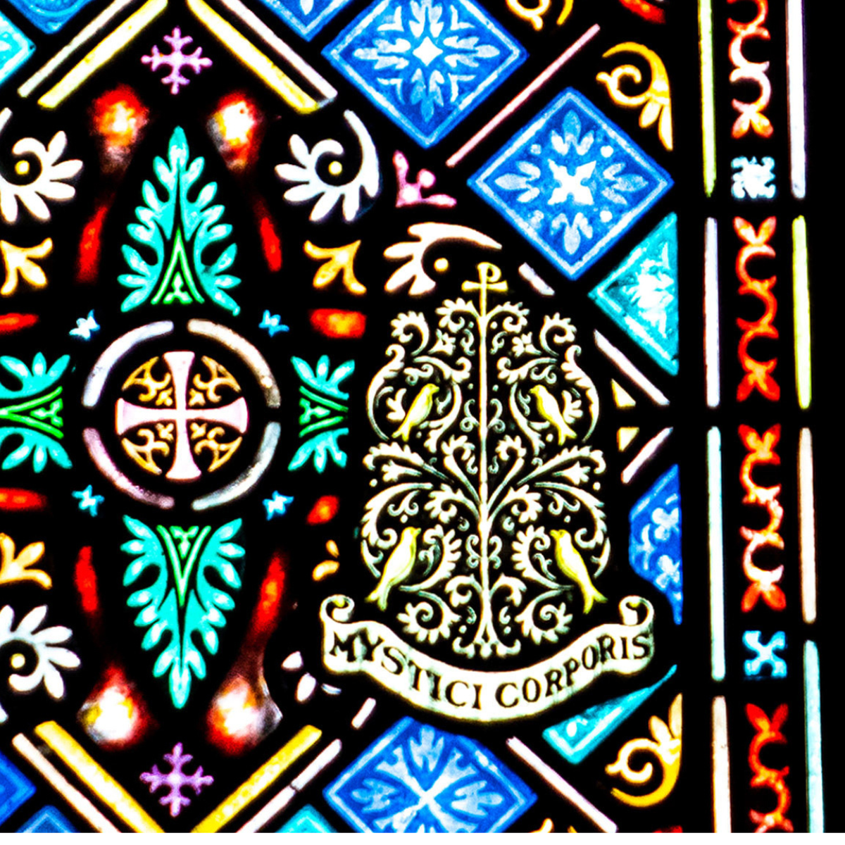 Colorful stained glass window featuring ornate geometric and floral patterns, with a leafy design and a banner below reading "MYSTICI CORPORIS" in gold letters.
