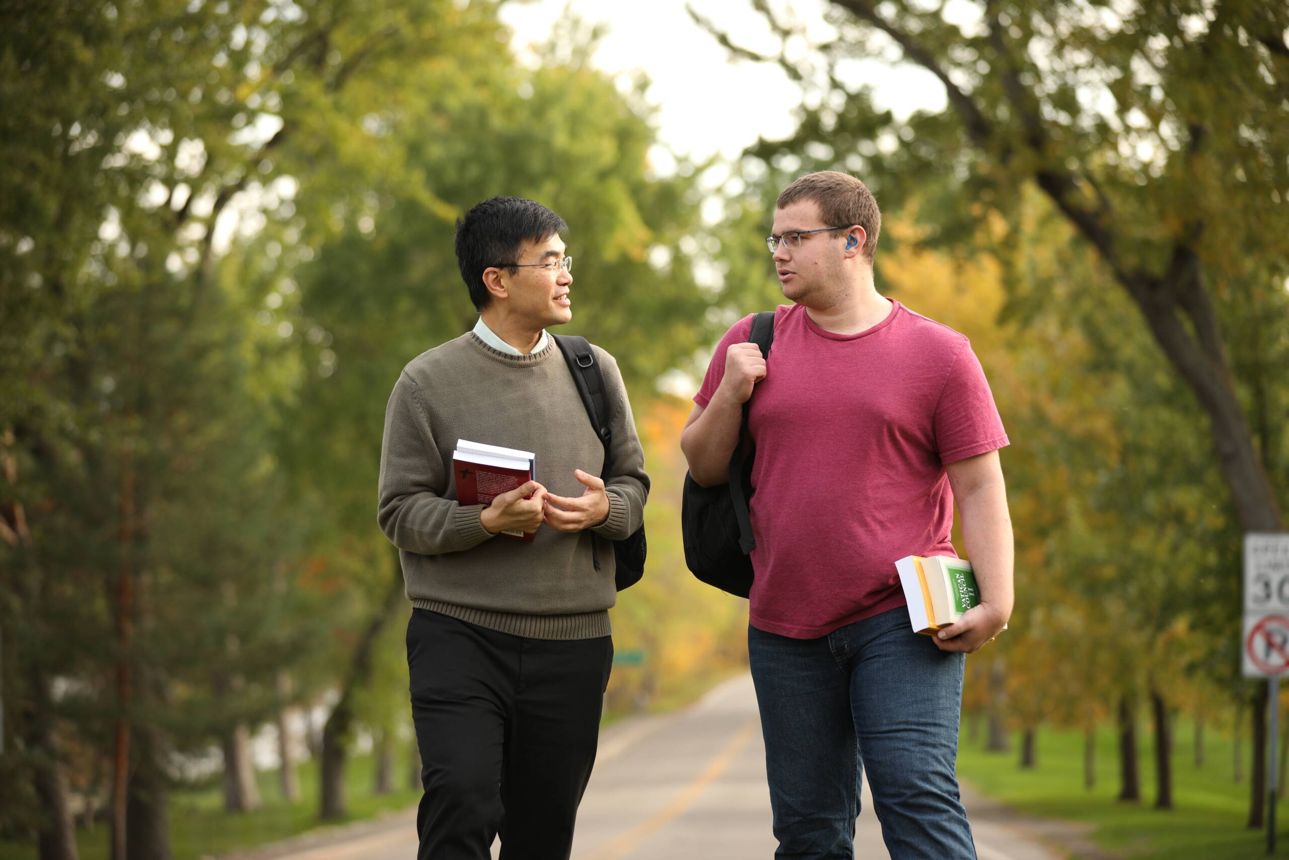 Two men walking and talking on a tree-lined road, each carrying books and backpacks. One wears a sweater and collared shirt; the other wears a red t-shirt and glasses. It's a bright, outdoor setting with greenery.