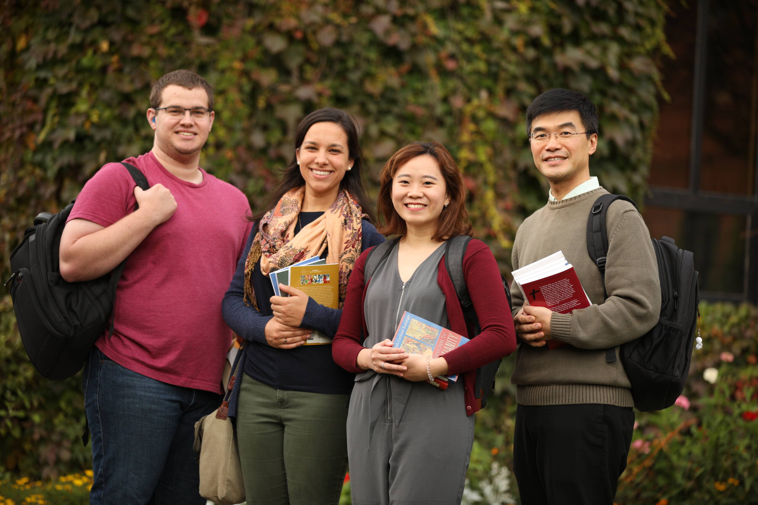 Four smiling adults stand outdoors in front of leafy greenery, holding books and wearing backpacks, suggesting they are students or colleagues.