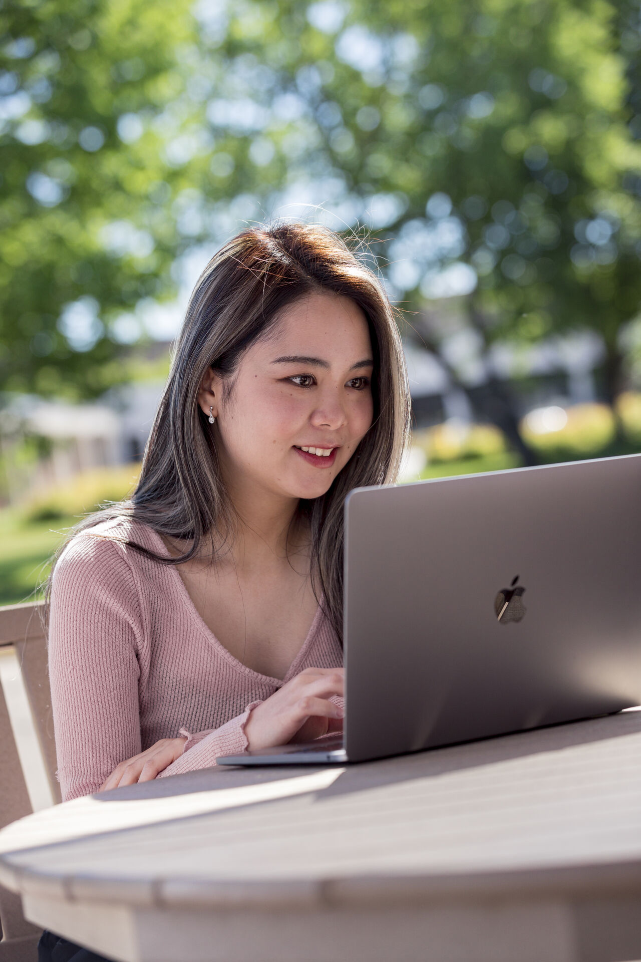 A woman with long hair sits at an outdoor table, smiling and working on a laptop. Trees and blurred greenery are visible in the sunlit background.