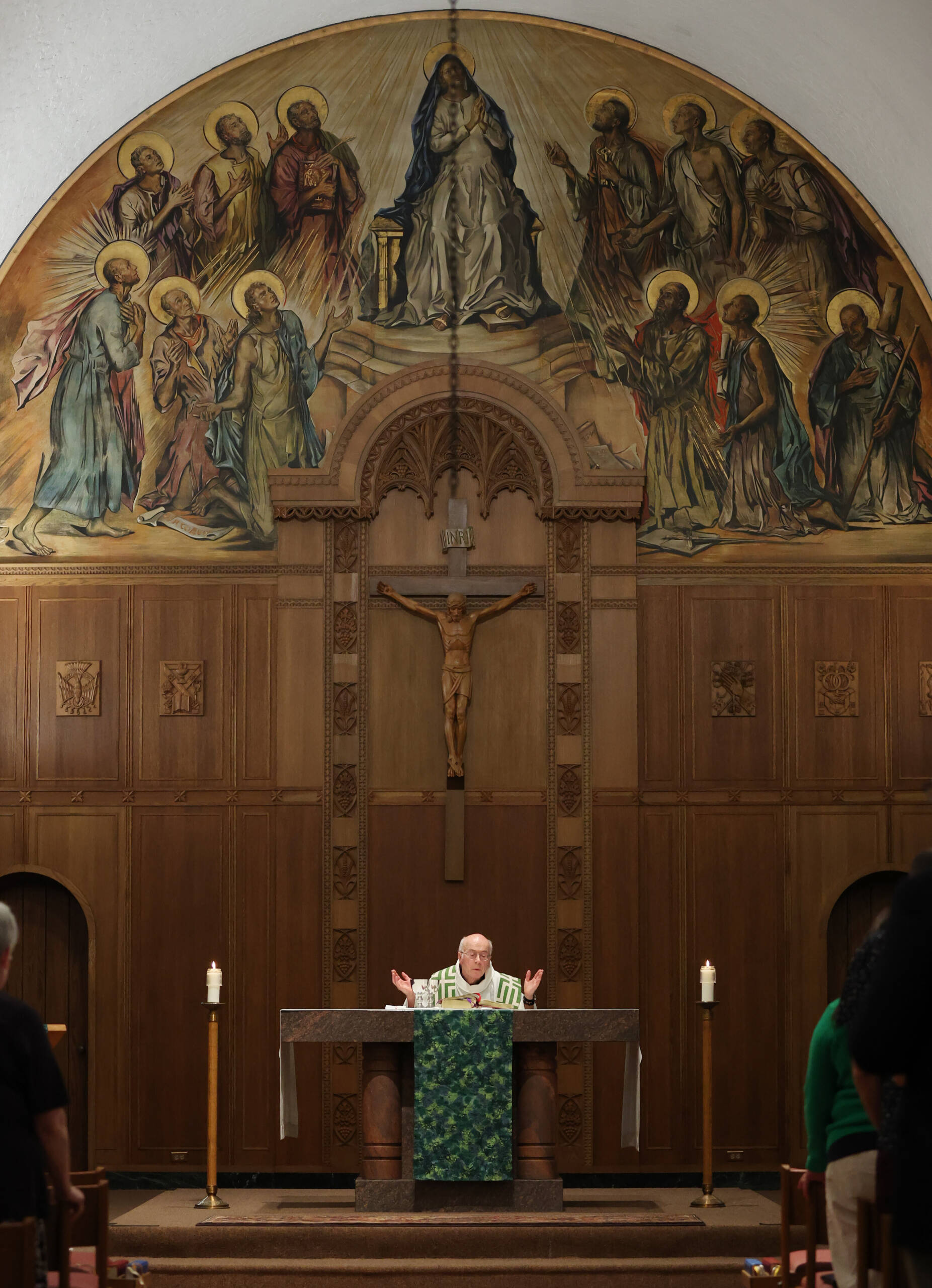 A priest stands at an altar with arms raised inside a church. Behind him are a crucifix and a large mural of religious figures, including one central figure seated under an arch, with candles on either side of the altar.