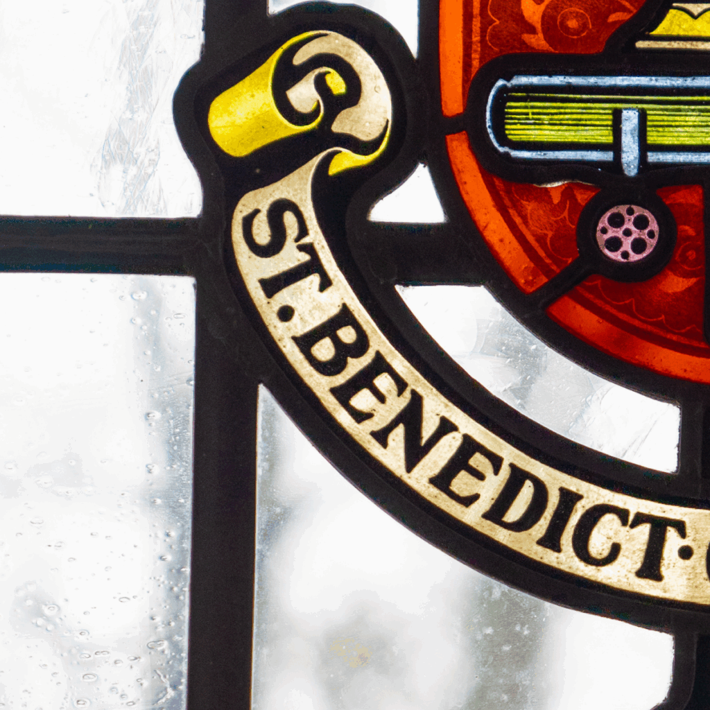 A stained glass window featuring the words "ST. BENEDICT" in gold letters on a black banner, with part of a shield and books visible above the text.