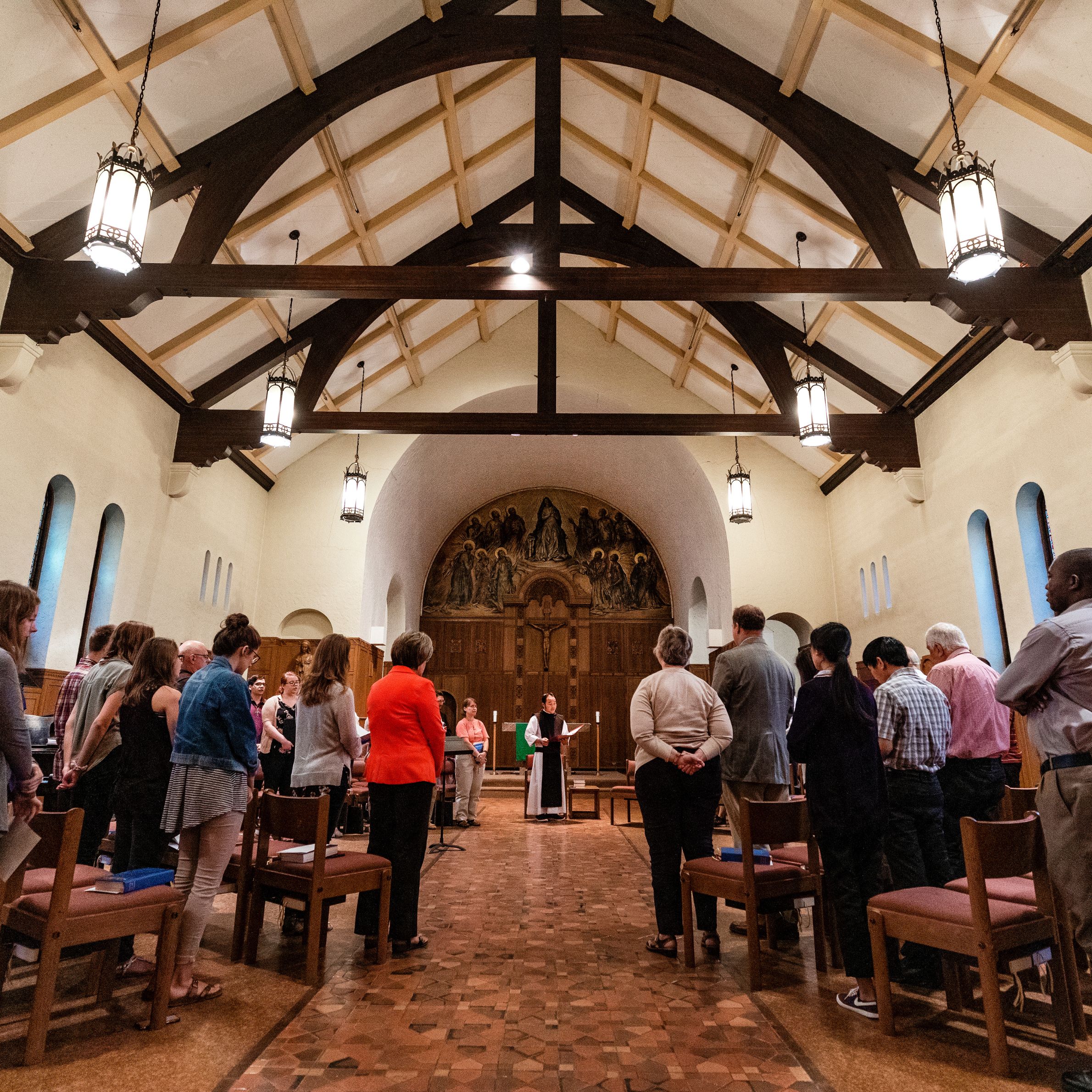 A group of people stands in a semicircle inside a church with vaulted ceilings and hanging lights, facing a person speaking at the front near the altar.