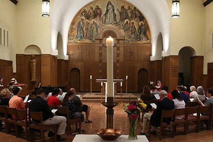 A group of people sit in a chapel, facing an altar with a lit candle. The chapel has wooden pews, a high ceiling, and a decorative mural above the altar depicting religious figures. Flowers and a bowl are placed near the altar.