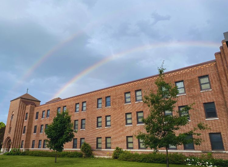 A double rainbow arcs across a cloudy sky above a large, brick building with multiple windows. Green grass, bushes, and trees are in front of the building.