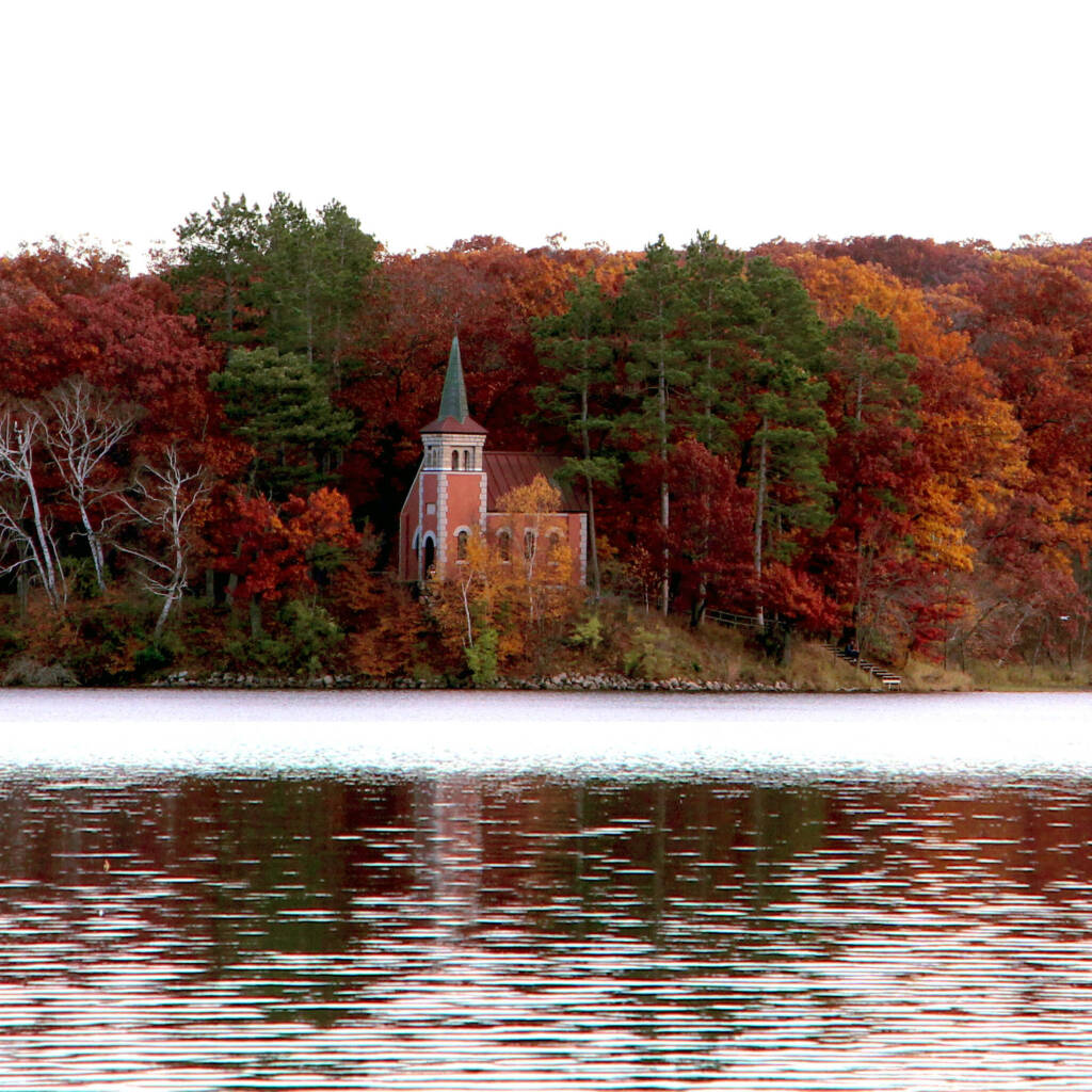 A small brick church with a steeple stands surrounded by colorful autumn trees on the shore of a calm lake, with the foliage reflecting in the water under an overcast sky.