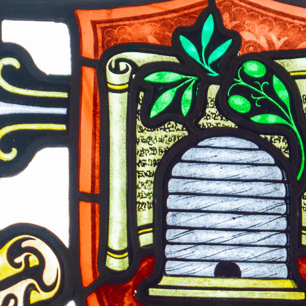 Stained glass artwork of a beehive with green leaves above it, set against a decorative background with scroll-like patterns and script.