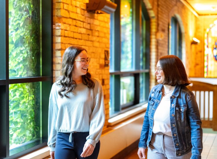 Two women are walking indoors by large windows with greenery outside, smiling and talking to each other. The hallway has exposed brick walls and warm lighting.