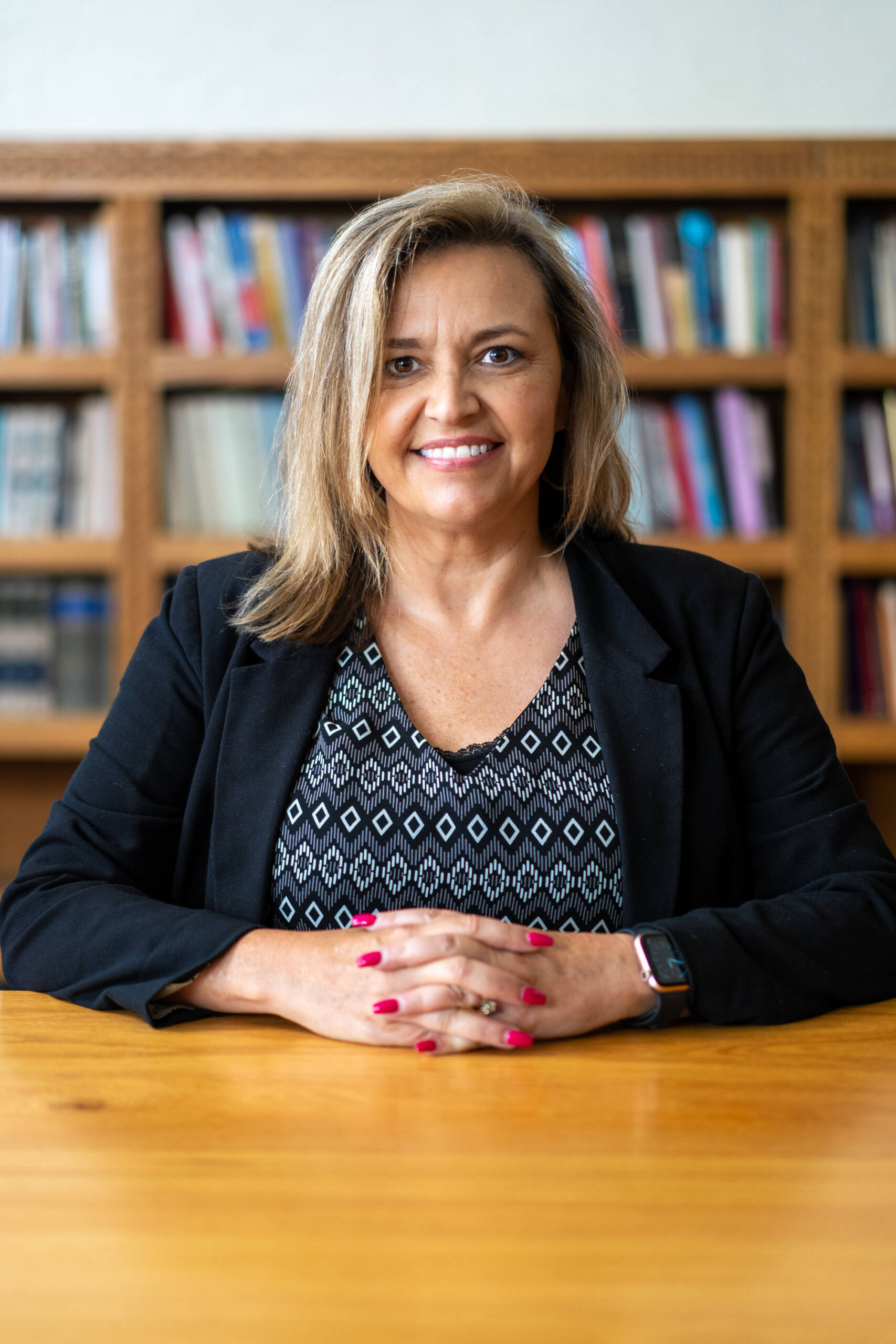 A woman with shoulder-length blonde hair sits at a wooden table, smiling. She wears a black blazer, patterned top, and a smartwatch. Bookshelves filled with books are blurred in the background.