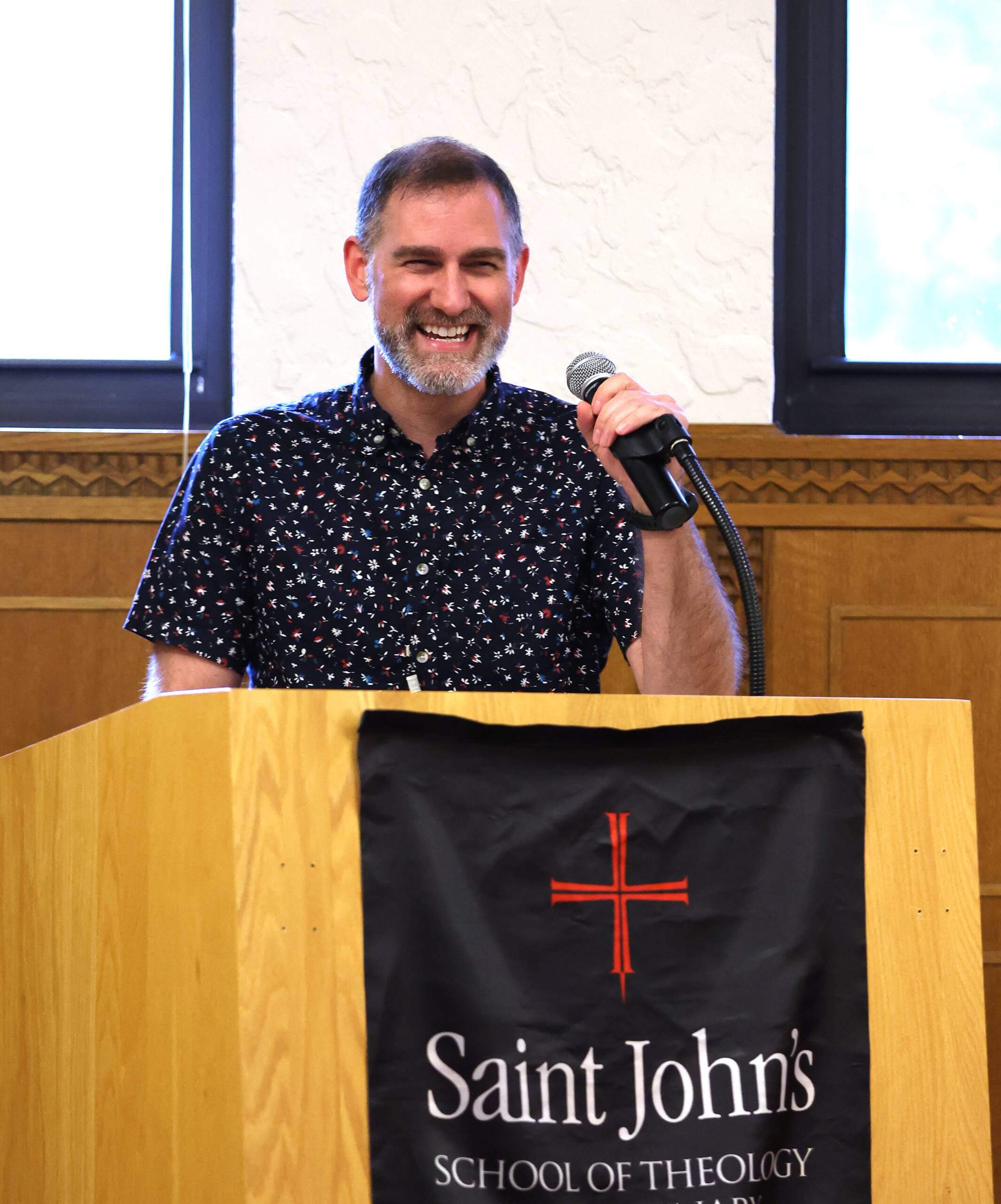 A smiling man in a patterned shirt stands at a wooden podium with a microphone. The podium has a black banner reading "Saint John's School of Theology." Two windows and a wood-paneled wall are in the background.
