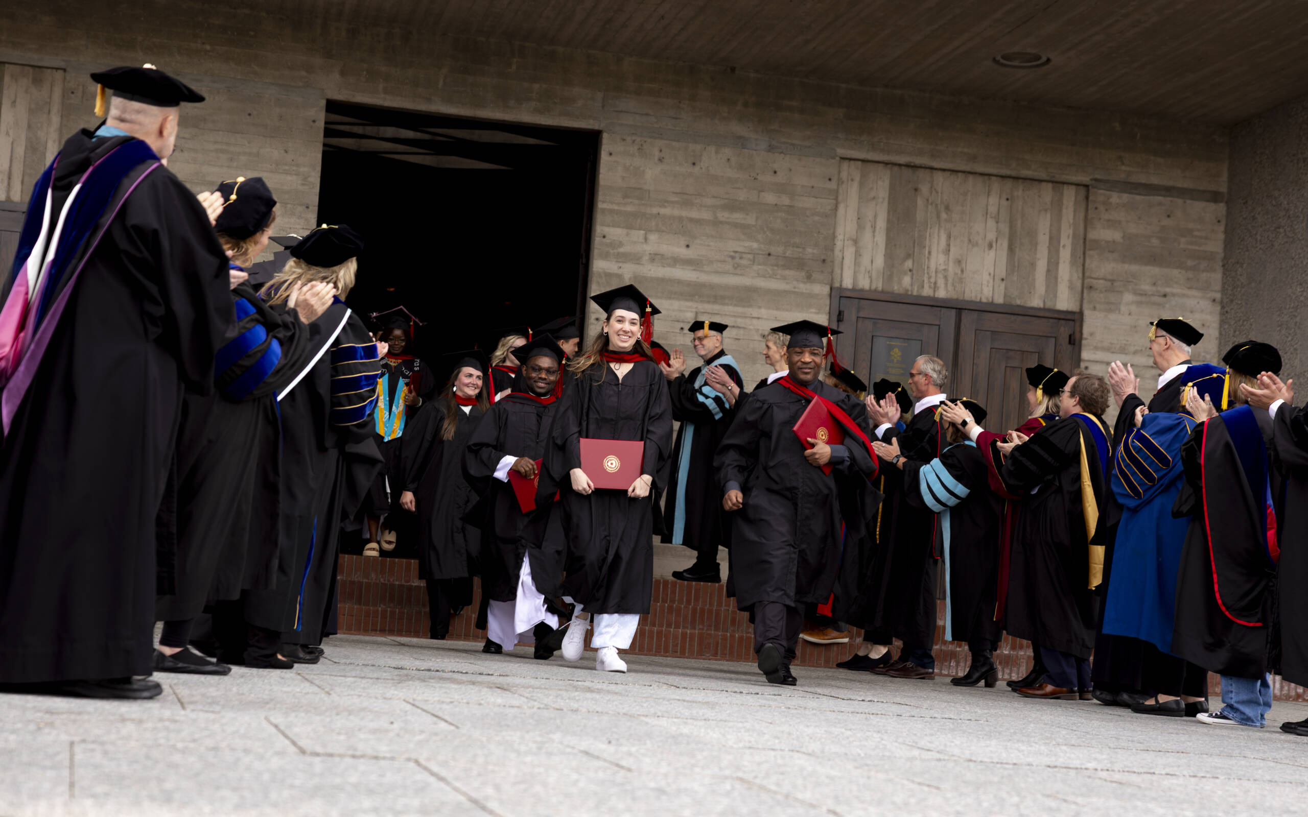 Graduates in caps and gowns walk out of a building holding diplomas while faculty in academic regalia stand in two lines, applauding and celebrating their achievement.