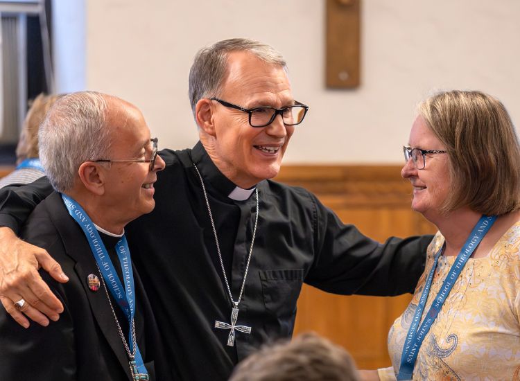 Three smiling adults, two men wearing clerical collars and a woman with glasses, stand close together indoors. The men have their arms around each other, and all wear blue lanyards. They appear to be in friendly conversation.