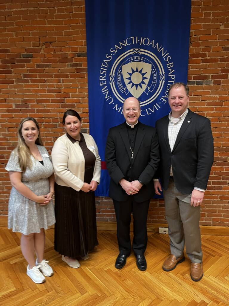 Four adults stand smiling in front of a blue university banner with a brick wall backdrop. Three are wearing business or semi-formal attire, while one wears clerical clothing. The group is standing on a polished wood floor.