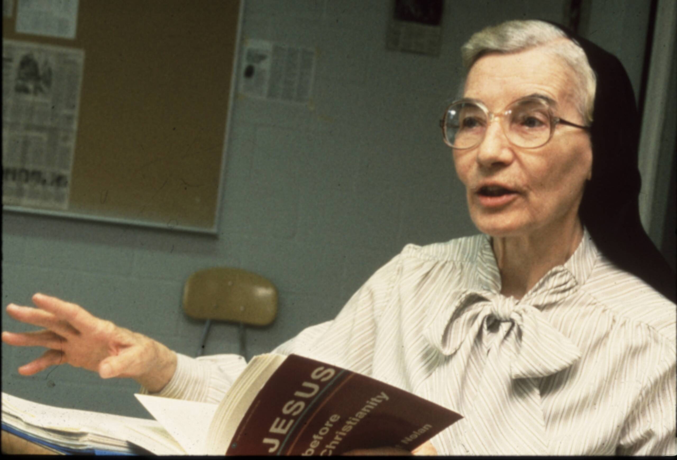 An elderly woman in glasses and a striped blouse with a bow, wearing a black headscarf, gestures while speaking. She is holding an open book titled "JESUS" in a classroom with a bulletin board behind her.