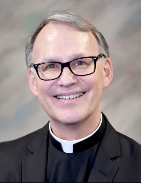 A man with short gray hair and black glasses, smiling, wears a black clerical suit with a white collar. The background is blurred in neutral tones.