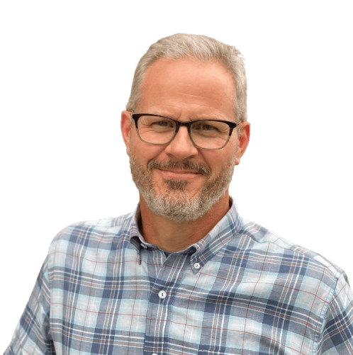 A middle-aged man with short gray hair, a beard, and glasses is wearing a blue plaid shirt and smiling at the camera against a plain light background.