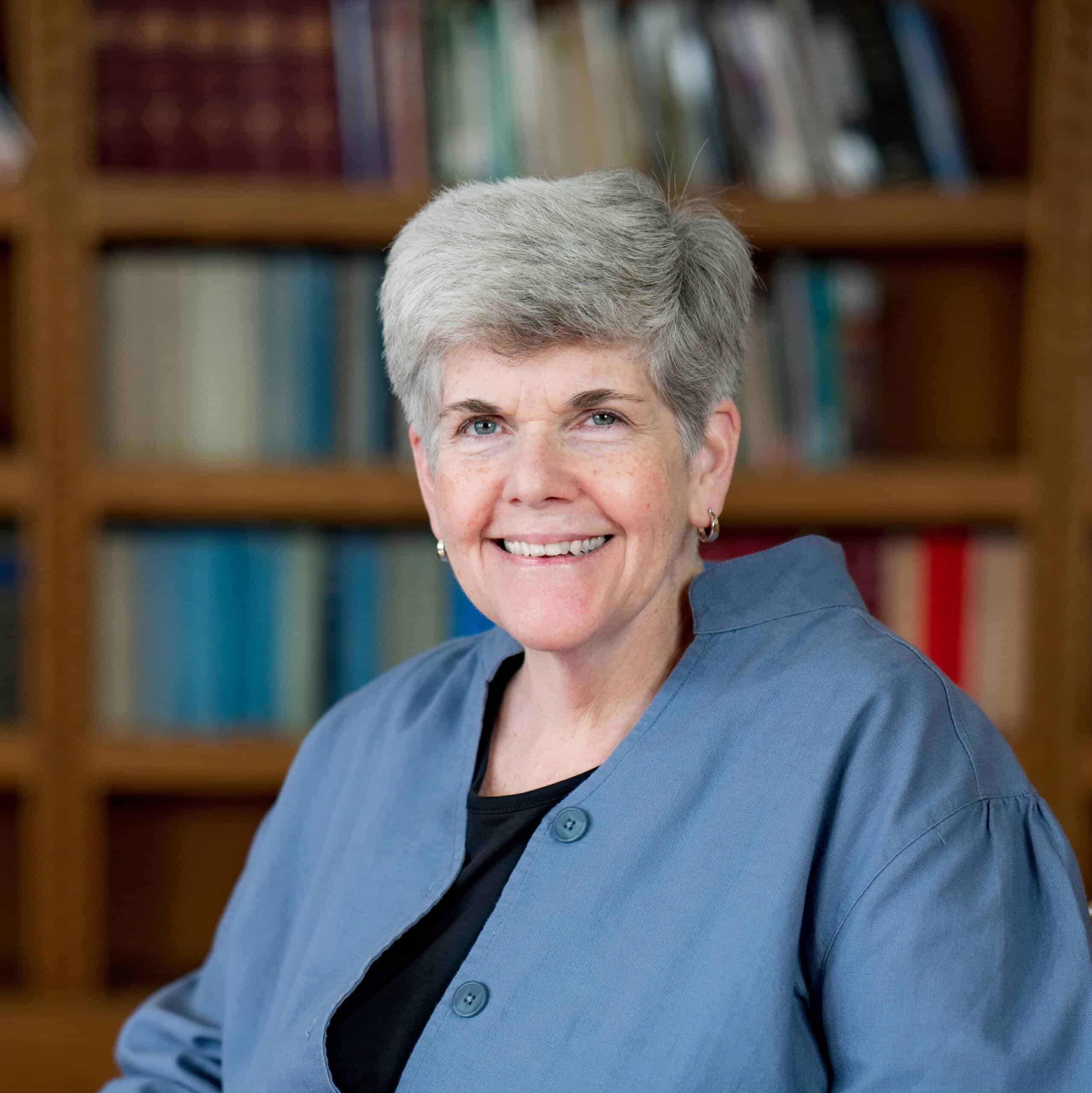 A smiling older woman with short gray hair, wearing a blue jacket over a black top, sits in front of a bookshelf filled with assorted books.