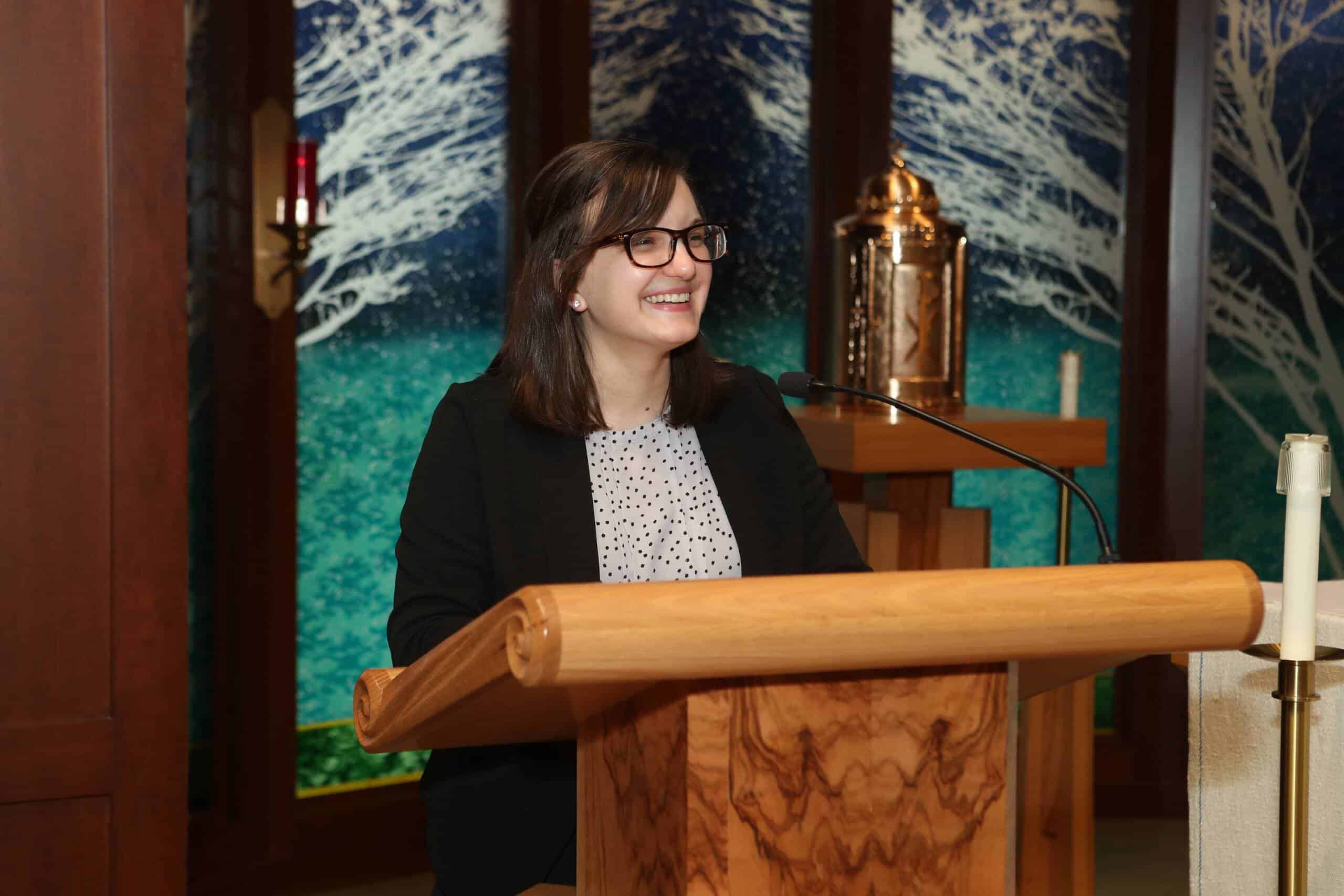 A woman with glasses and dark hair, dressed in a black blazer and white blouse, smiles while speaking at a wooden podium in a room with decorative blue-green art and a gold religious object in the background.