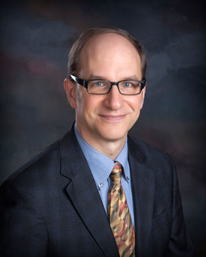 A man with short brown hair and glasses is smiling. He is wearing a dark suit, a light blue shirt, and a patterned tie. The background is a gradient of dark colors.