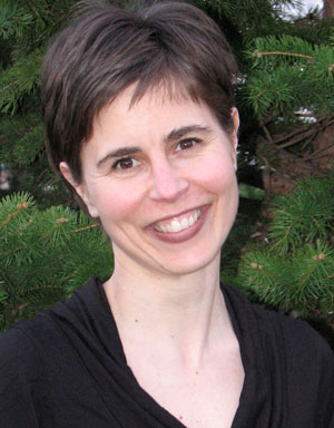 A smiling woman with short, dark hair stands in front of a backdrop of green pine trees. She is wearing a black top and is looking directly at the camera.