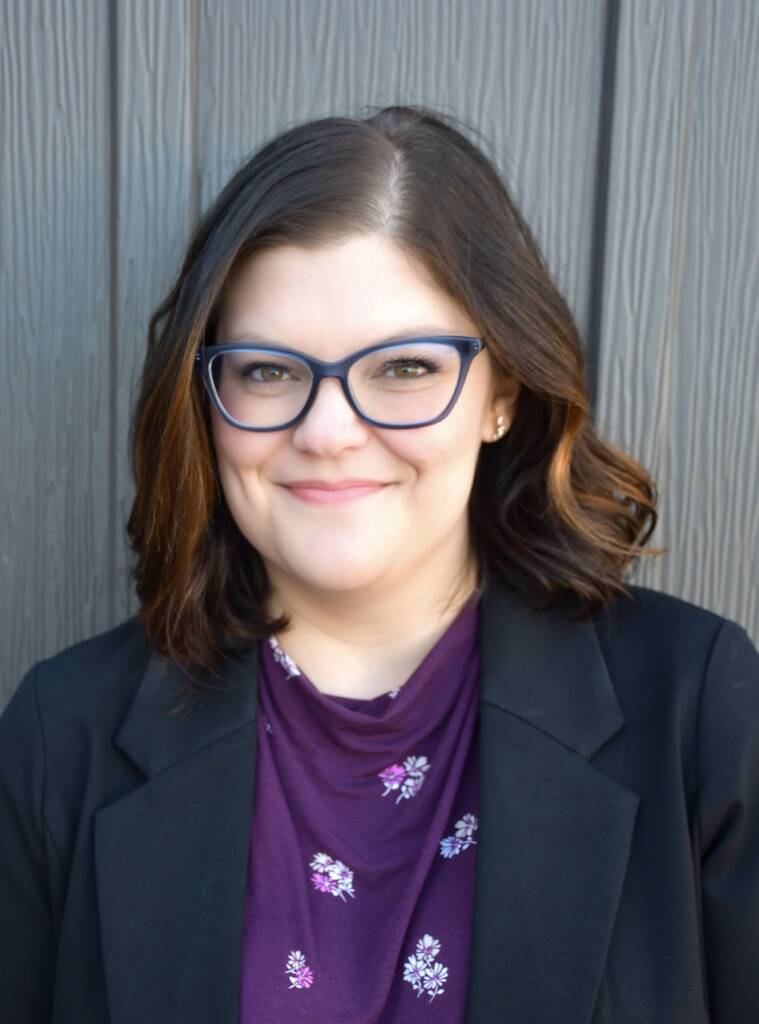 A person with shoulder-length brown hair, wearing blue-rimmed glasses, a dark blazer, and a purple top with floral patterns is seen standing in front of a vertically paneled wooden wall, smiling at the camera.